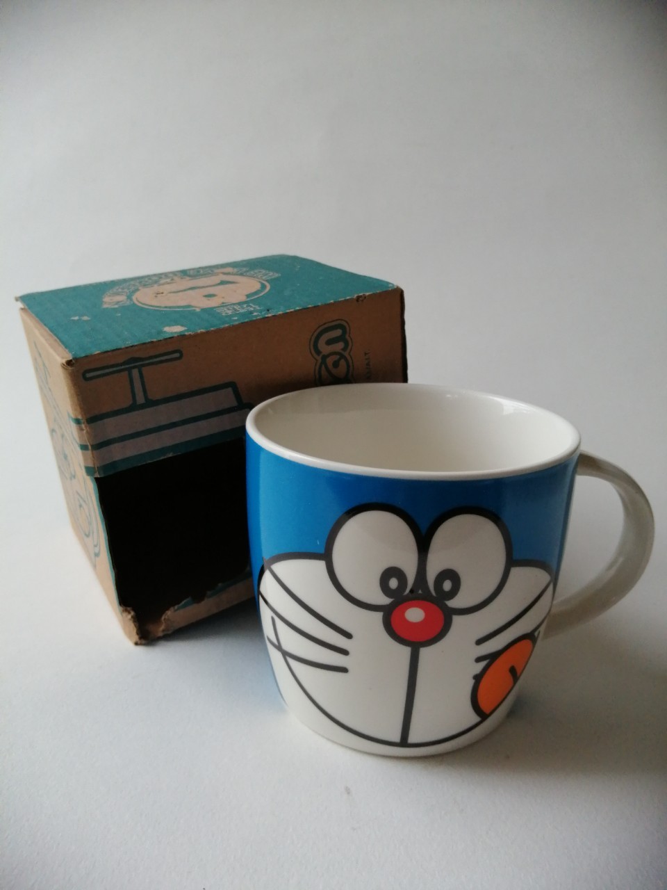 แก้วน้ำเซรามิก "แก้วโดราเอม่อน หน้ายิ้มแลบลิ้นด้านข้าง **ไม่มีฝายางสีน้ำเงิน พร้อมกล่อง" ของสะสมจากเทสโก้ โลตัส***สภาพสะสมเก่าเก็บ กล่องมีตำหนิตามภาพ